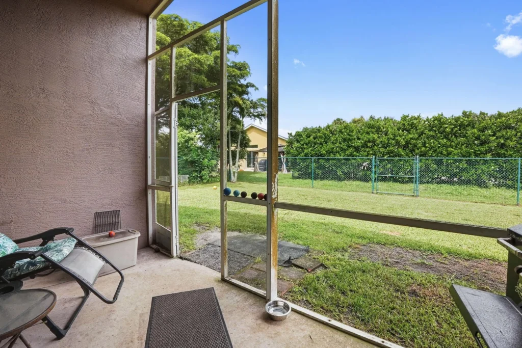 20260924162019116982000000-o - Lake Worth Homes - Lake Worth Homes Covered patio with open screen door looking onto a grassy backyard and a green fence; croquet balls lined along the door frame.