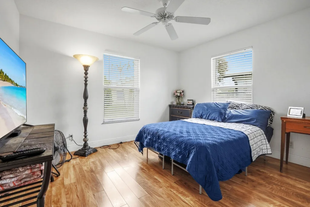 20260924161926643657000000-o - Lake Worth Homes - Lake Worth Homes Bright bedroom with a blue quilted bed, two matching pillows, and a zebra-patterned accent on the pillows; hardwood floor, two windows with blinds, and a ceiling fan. A tall floor lamp sits beside the window, and a TV on a stand is opposite the bed.