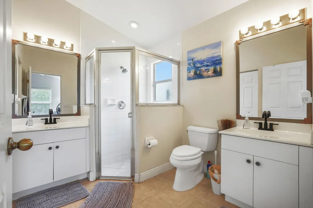 20260924161954081444000000-o - Lake Worth Homes - Lake Worth Homes Bright bathroom with dual sinks, white vanities, framed mirrors, and a glass-enclosed shower beside a toilet.