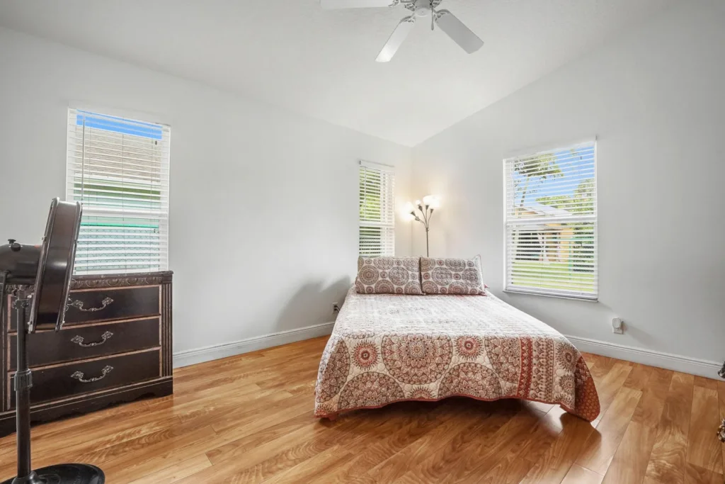 20260924161939996007000000-o - Lake Worth Homes - Lake Worth Homes Bright bedroom with a bed dressed in a red-patterned quilt and two pillows, wooden dresser on the left, floor fan nearby, and large windows with blinds.