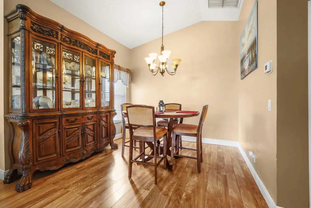 20260924161803274670000000-o - Lake Worth Homes - Lake Worth Homes Traditional dining room with a large ornate wooden china cabinet on the left, a round table and four upholstered chairs, and a chandelier overhead on warm beige walls.