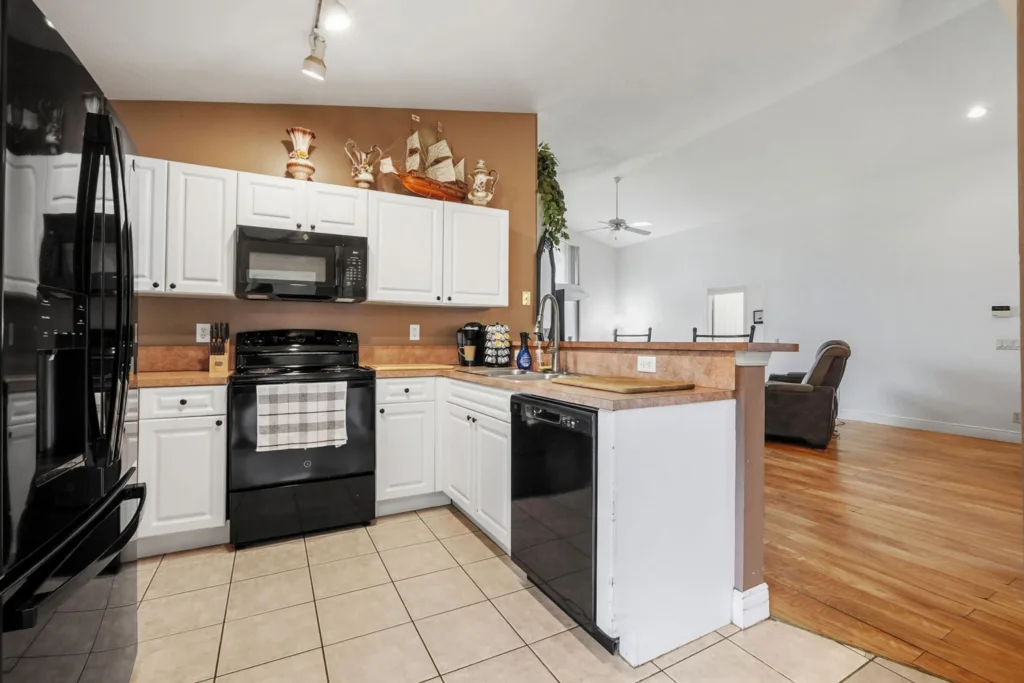 20260924161836916632000000-o - Lake Worth Homes - Lake Worth Homes Kitchen with white cabinets, black appliances (stove, microwave, fridge), and beige tile floor; brown accent wall above the counters with decorative items on top.
