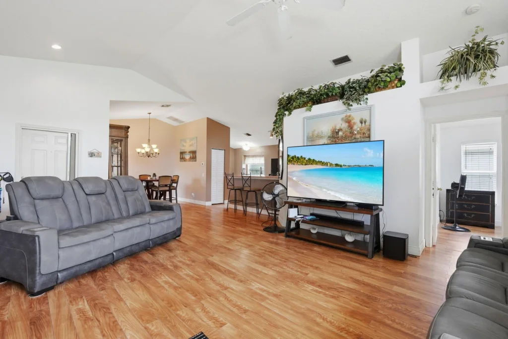 20260924161756414477000000-o - Lake Worth Homes - Lake Worth Homes Bright living room with hardwood flooring, a large gray sectional sofa, and a TV displaying a tropical beach scene on a stand beneath decorative greenery.