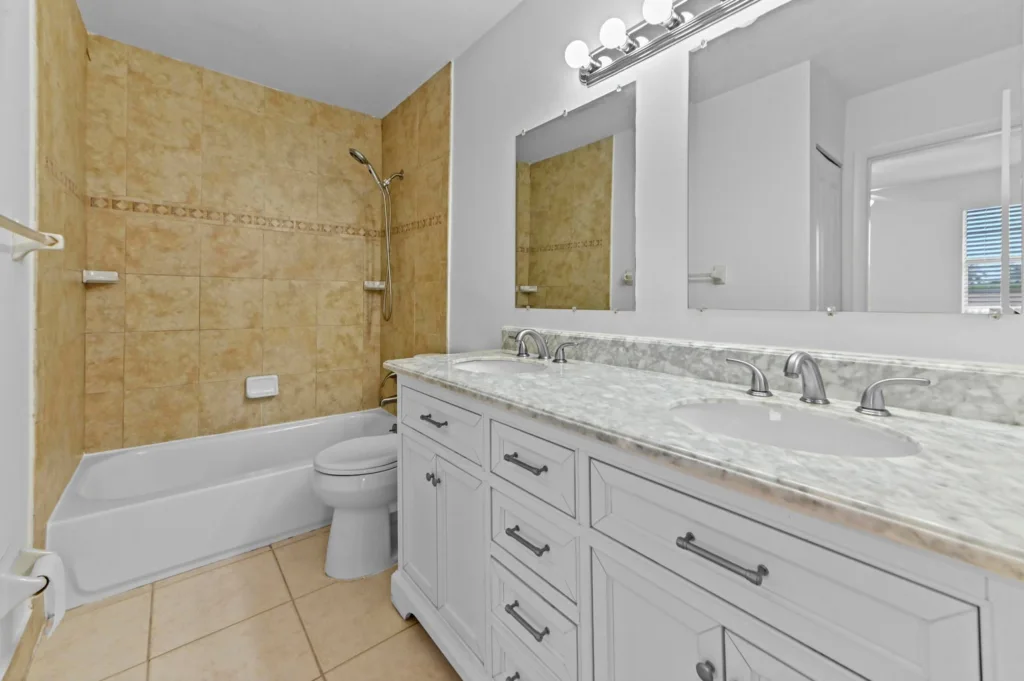 20260930184856122602000000-o - Lake Worth Homes - Lake Worth Homes Bathroom with beige tiled shower and tub, white toilet, and a long marble countertop with double sinks under bright vanity lights.