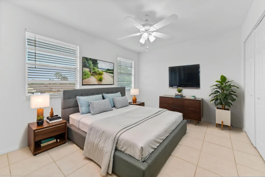 20260930184825775869000000-o - Lake Worth Homes - Lake Worth Homes Modern bedroom with a gray upholstered king bed, two nightstands, lamps, and a dresser with a wall-mounted TV.
