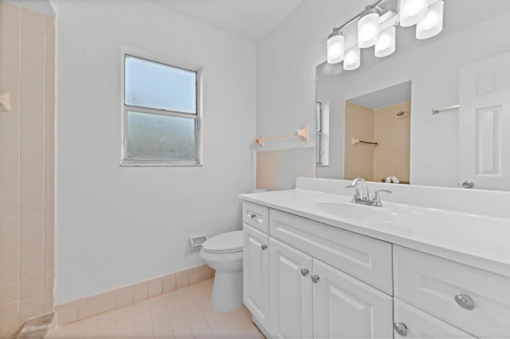 20260930184652607161000000-o - Lake Worth Homes - Lake Worth Homes White bathroom with a single-sink vanity, chrome faucet, large mirror, and frosted window; beige floor tiles and shower area visible in the corner.