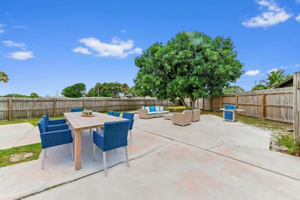 20260930185116081725000000-o - Lake Worth Homes - Lake Worth Homes Outdoor backyard scene with a large green tree, blue patio chairs around a wooden dining table, and a gray wicker seating area on a concrete patio behind a wooden fence.