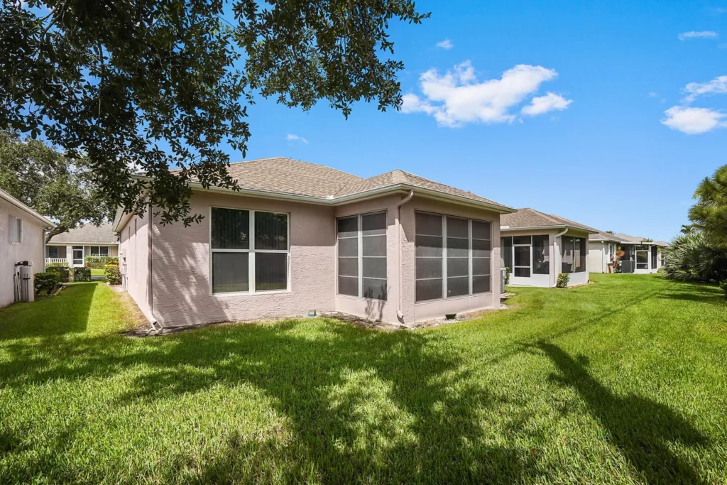 Backyard of a beige single-story house with a screened-in patio and lush green lawn under a bright blue sky.