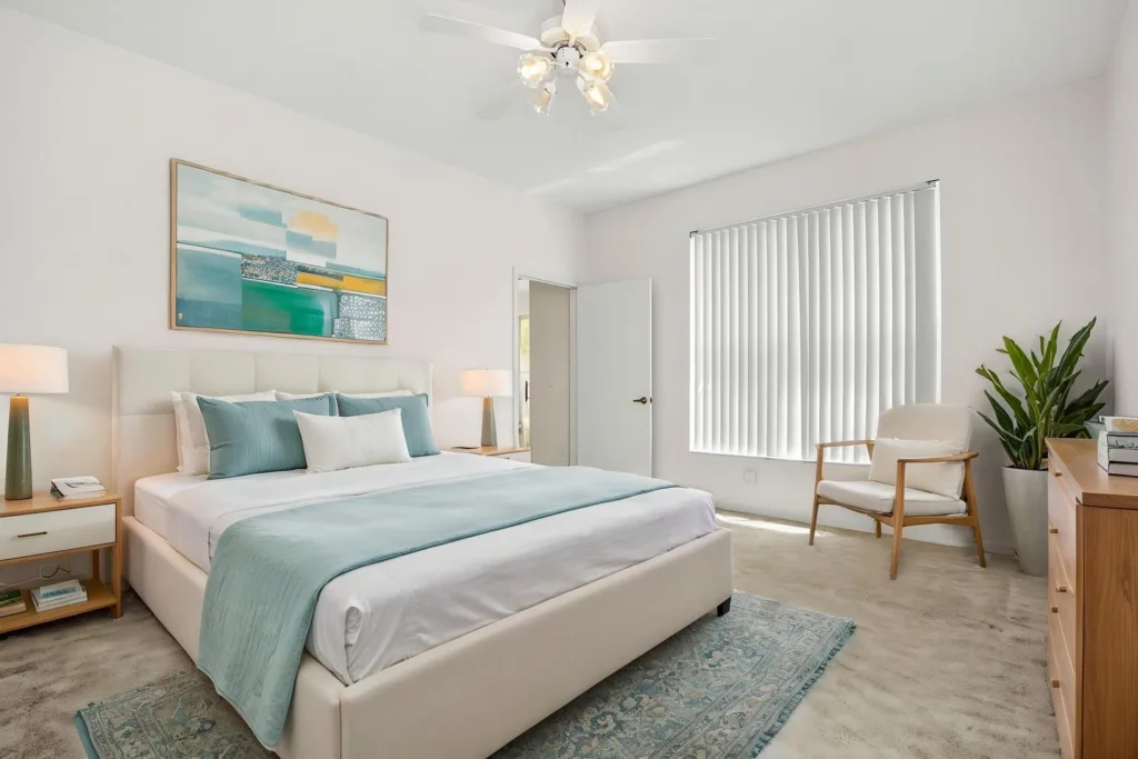 Bright white bedroom with a plush upholstered bed, teal accents, two nightstands, and a window with vertical blinds.