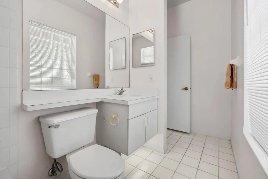 White bathroom with a long vanity, large mirror, and toilet; window blinds reflect light from the right. A beige towel hangs by the door.