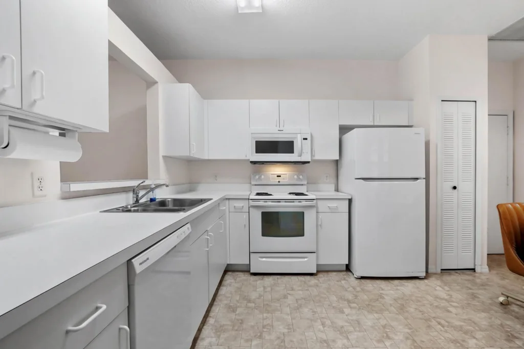Bright all-white kitchen with upper and lower cabinets, white countertops, a double sink, and white appliances (dishwasher, stove with microwave, and a refrigerator) along beige-tiled floor.