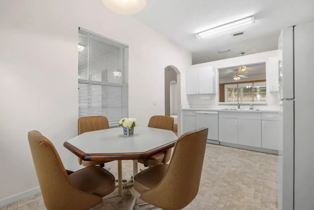 Dining area with a white round table and four tan upholstered chairs near a large window with blinds.|Rooms are light and clean with neutral tones and a small flower centerpiece on the table.