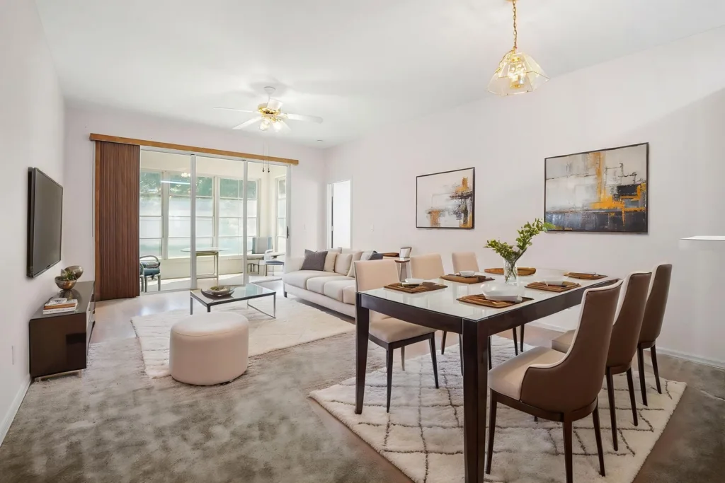 Open-concept living and dining room with a glass dining table, eight beige chairs, and abstract wall art on pale walls.