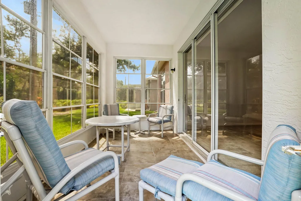 Sunlit enclosed patio with blue striped cushioned chairs around a white round table, glass walls showing a green yard outside.