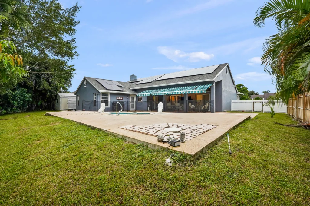 20261001130759458693000000-o - Lake Worth Homes - Lake Worth Homes Backyard of a modern house with a rectangular in-ground pool, screened enclosure, and a shaded patio under a striped awning.