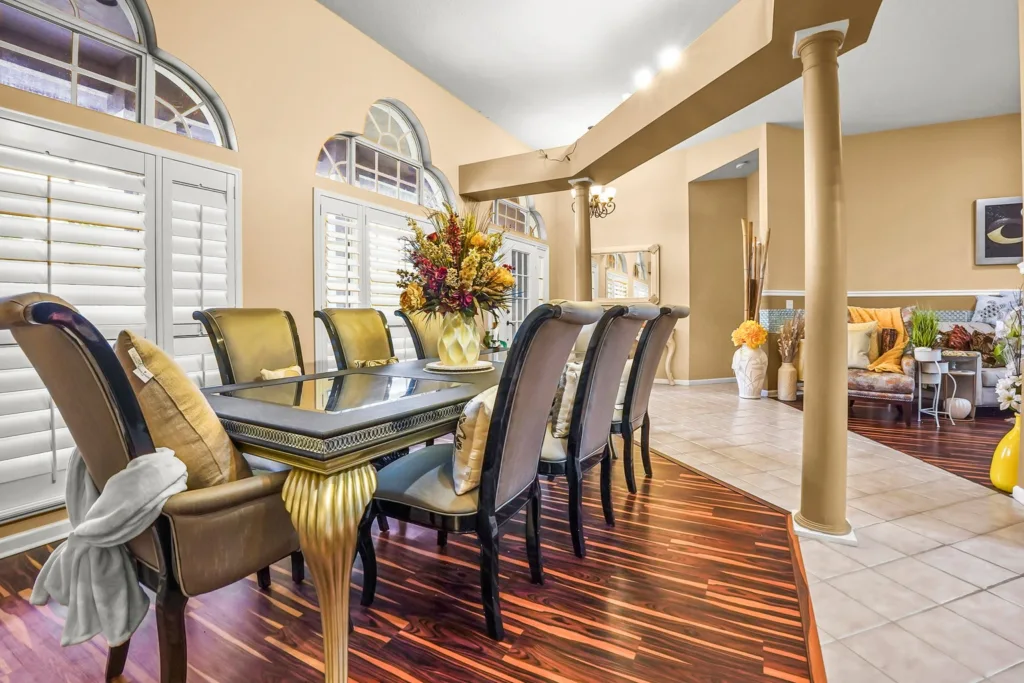 20261001125850574990000000-o - Lake Worth Homes - Lake Worth Homes Elegant dining room with a glass-top table, gold-upholstered chairs, and a colorful floral centerpiece under arched windows with shutters.