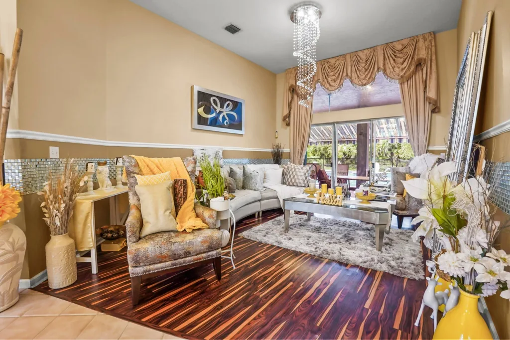 20261001125804421423000000-o - Lake Worth Homes - Lake Worth Homes Cozy living room with beige walls, a chandelier, and a gray sectional facing a glass coffee table on a striped wood floor.