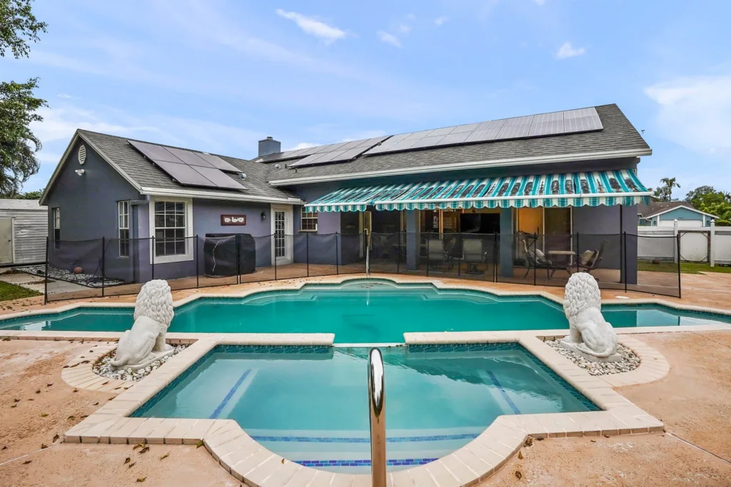 20261001130640384535000000-o - Lake Worth Homes - Lake Worth Homes Backyard of a gray house with solar panels, a fenced pool, striped green awning, and two stone lion statues at the pool edges.