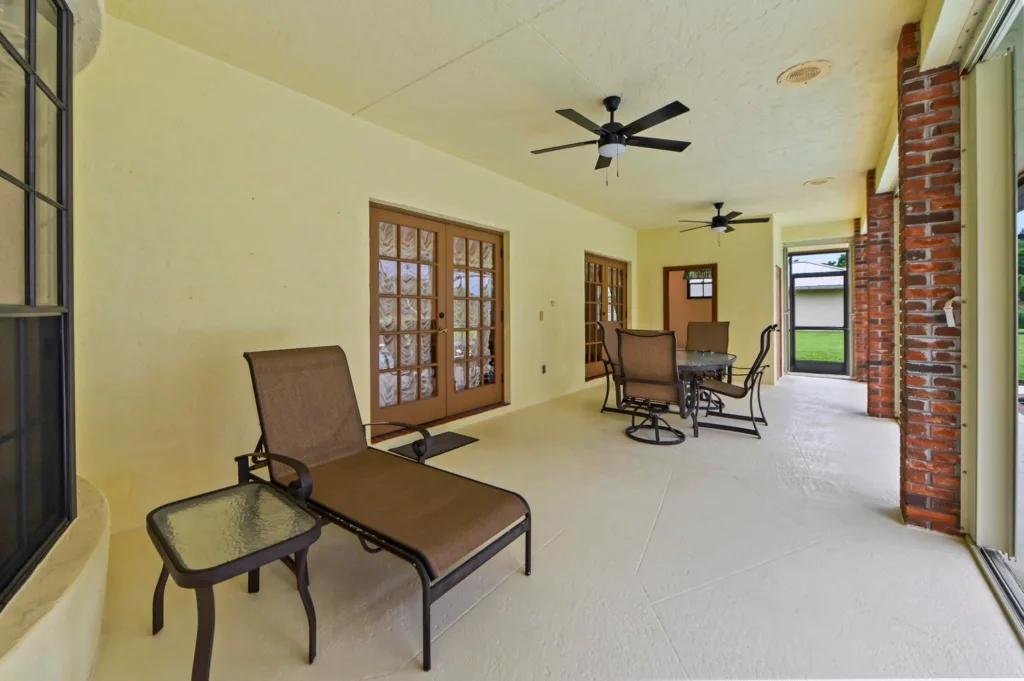 20260930221644222456000000-o - Lake Worth Homes - Lake Worth Homes Covered patio with yellow walls, brick columns, ceiling fans, and outdoor furniture on a light concrete floor. A lounge chair and glass-side table are in the foreground, with French doors along the wall.