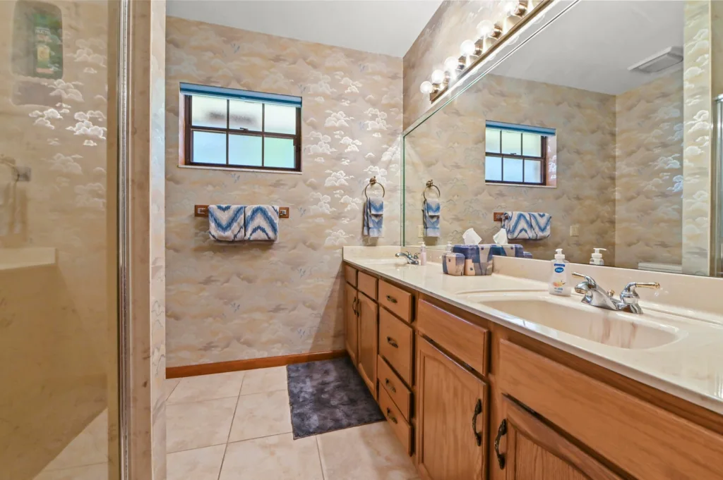 20260930221432140485000000-o - Lake Worth Homes - Lake Worth Homes Bathroom with a double-sink vanity, light wood cabinets, large mirror, and cloud-pattern wallpaper with blue towels.