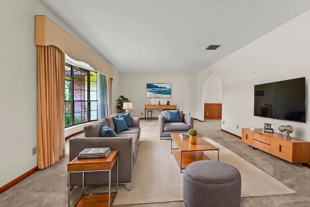 20260930222651225887000000-o - Lake Worth Homes - Lake Worth Homes Cozy mid-century living room with gray sofas, blue cushions, and a large window with peach curtains. A wooden coffee table and TV console are visible.