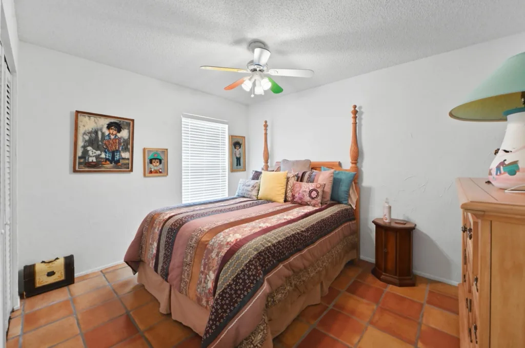 20260925125456209901000000-o - Lake Worth Homes - Lake Worth Homes Cozy bedroom with a wood four-poster bed, striped quilt, and colorful throw pillows; terracotta tile floor and a ceiling fan with colored blades overhead.