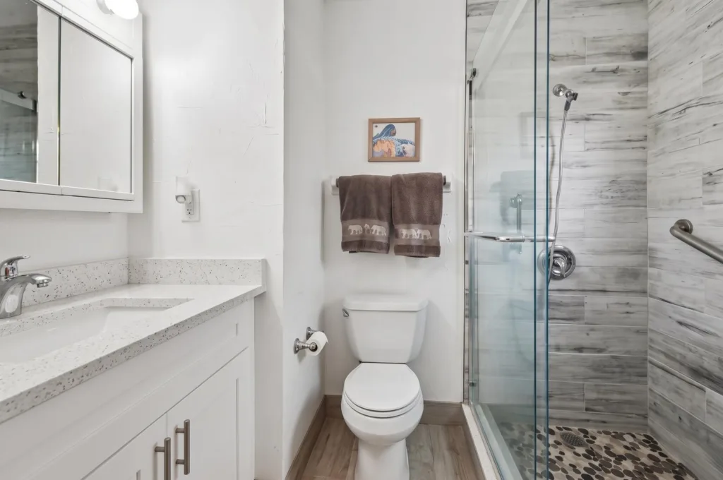 20260925161934643157000000-o - Lake Worth Homes - Lake Worth Homes White bathroom with vanity, mirror cabinet, toilet, brown towels on a rack, framed seaside artwork, and a glass shower with gray marble tiles.