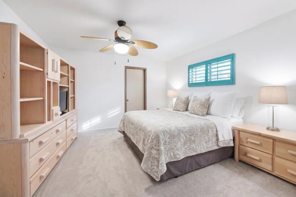 20260925162642669443000000-o - Lake Worth Homes - Lake Worth Homes Bright bedroom with a king bed, light wood dresser, built-in shelving, two nightstands, and teal window shutters.
