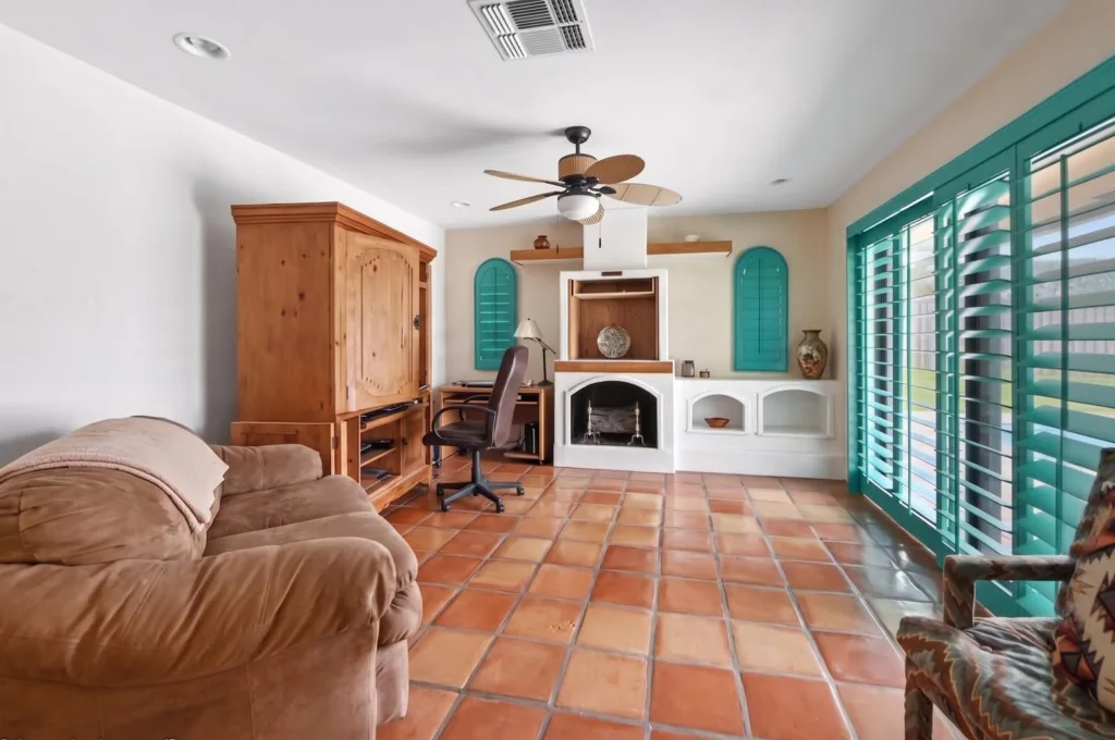 20260925162157439519000000-o - Lake Worth Homes - Lake Worth Homes Living room with a tan sofa, wooden armoire, desk, and a white fireplace flanked by teal shutters.