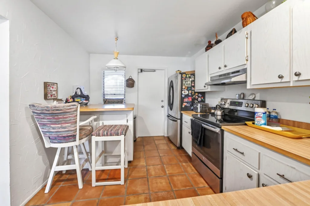 20260925125339286451000000-o - Lake Worth Homes - Lake Worth Homes Cozy white kitchen with a small breakfast bar and two striped upholstered stools, terracotta tile floor.