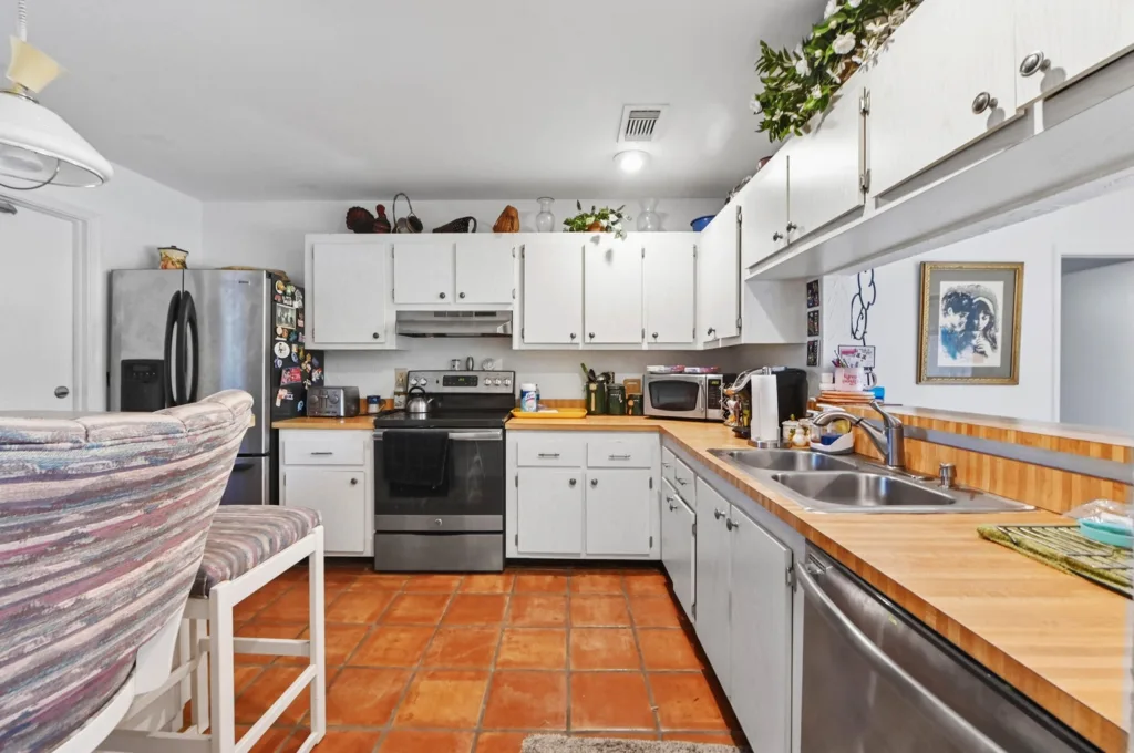 20260925125327722506000000-o - Lake Worth Homes - Lake Worth Homes Bright U-shaped kitchen with white cabinets, stainless fridge and stove, wooden countertops, and terracotta tile floor with potted plants on top.
