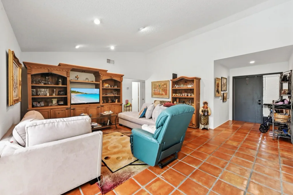 20260925125230251726000000-o - Lake Worth Homes - Lake Worth Homes Living room with terracotta tile floor, beige sofas, a teal recliner, and large wooden display cabinets with knickknacks and a TV.