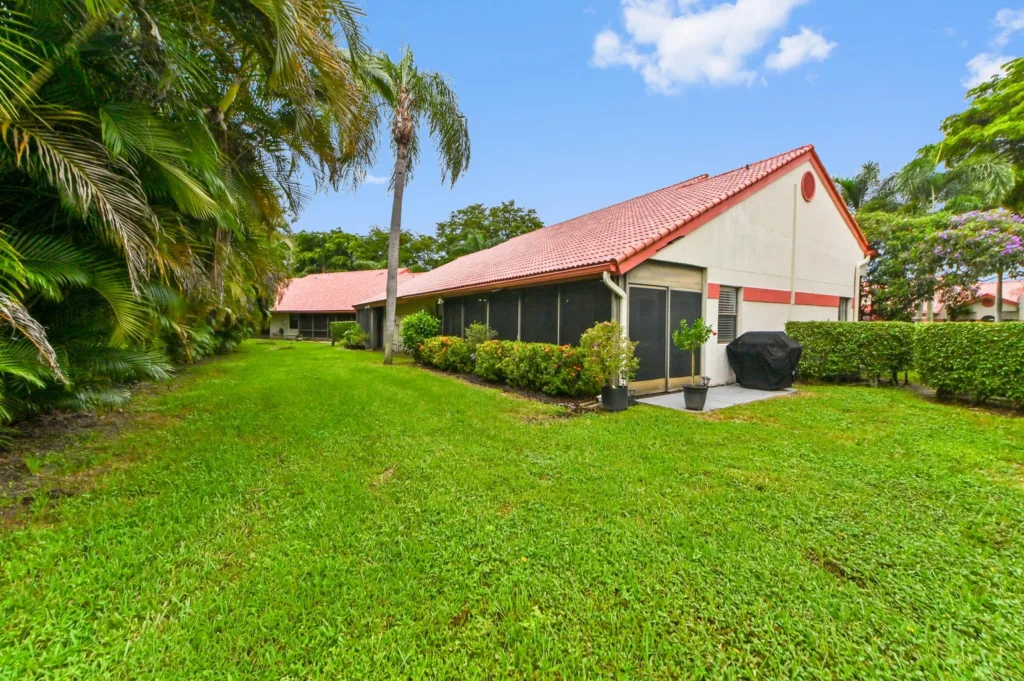 Suburban backyard with a red-tiled roof house, screened porch, green lawn, and palm trees along the edge.
