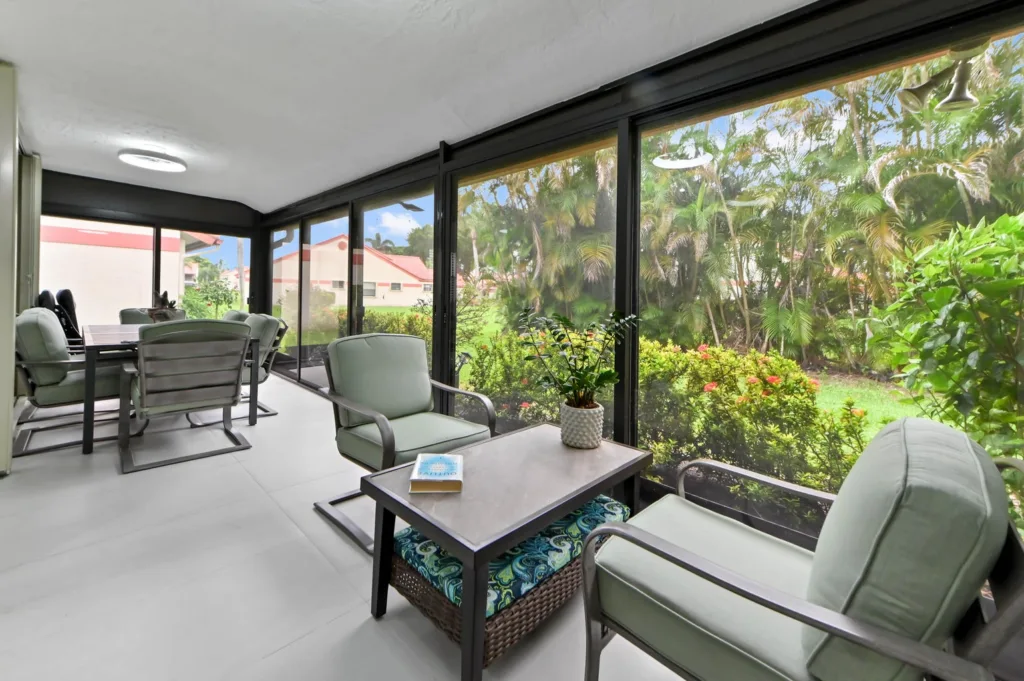 Sunroom with glass walls and mint-green cushioned chairs around a coffee table with a small plant, overlooking a lush garden.