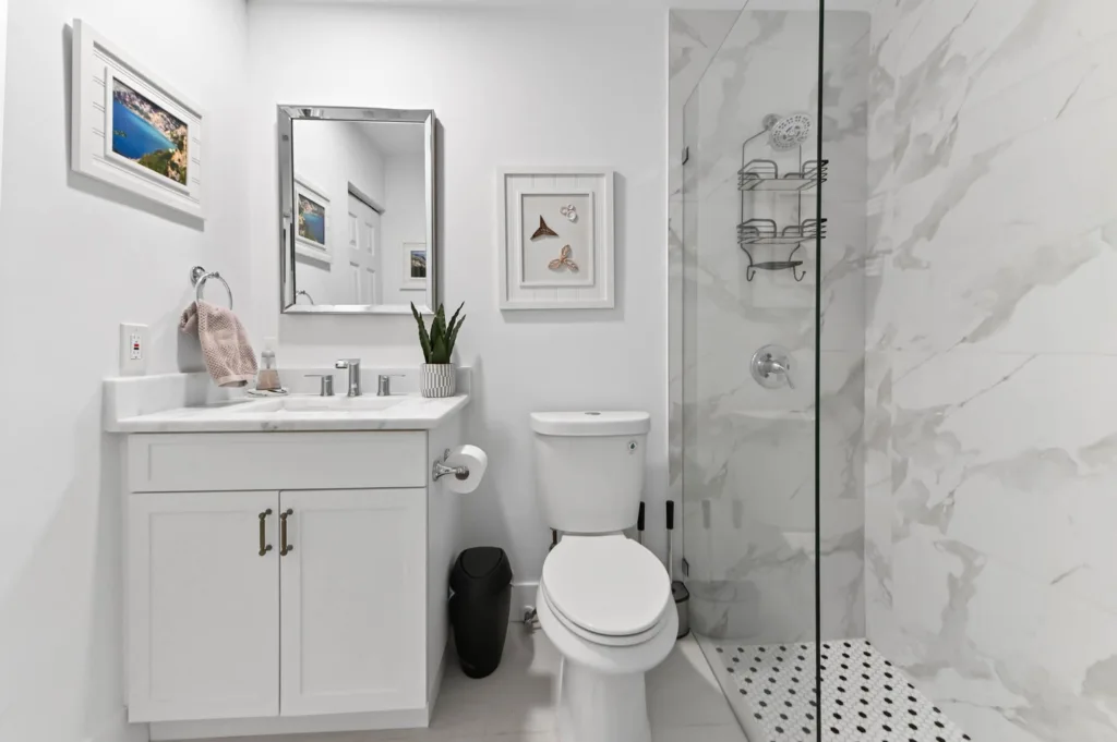 Bright, modern bathroom with white vanity, marble countertop, wall mirror, and framed artwork above the toilet area; glass-enclosed shower with marble walls in the background.