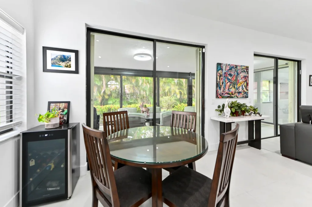 Bright dining area with a round glass table, four wooden chairs, and large sliding doors opening to a lush outdoor patio.
