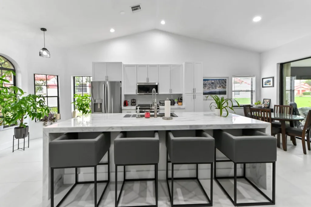Bright modern kitchen with a large marble island, six gray bar stools, white cabinets, and stainless steel appliances; dining area nearby with plants by windows.