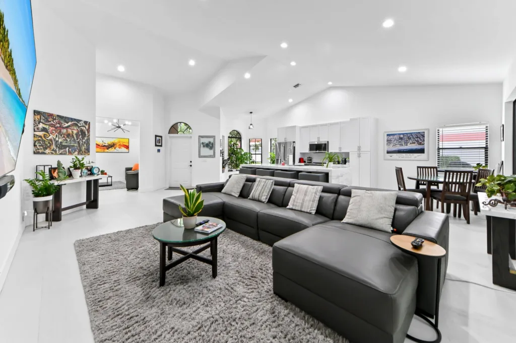 Open-concept living area with a gray leather sectional, white walls, and modern kitchen in the background.
