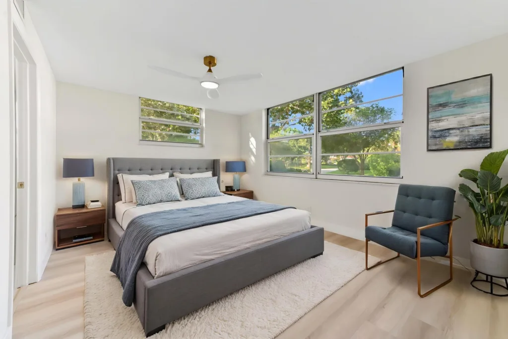 20261002165428159059000000-o - Lake Worth Homes - Lake Worth Homes Bright modern bedroom with a gray upholstered bed, matching nightstands, and large windows showing trees outside.