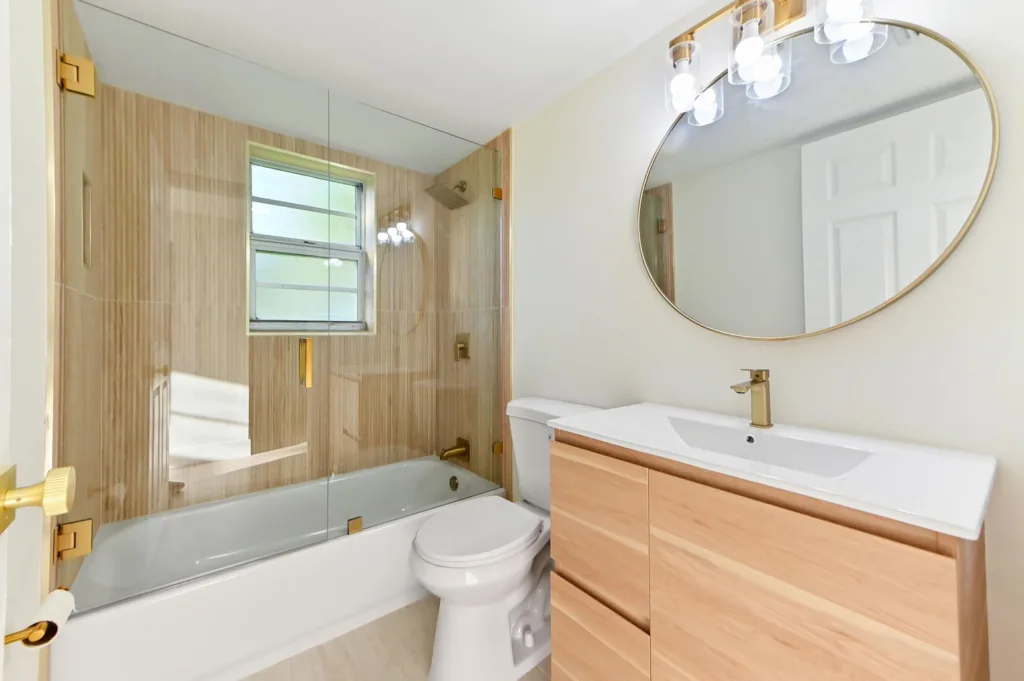 20260929180550409316000000-o - Lake Worth Homes - Lake Worth Homes Modern bathroom with a glass-enclosed shower, beige tiled walls, white tub, and a wooden vanity with a round mirror and gold fixtures