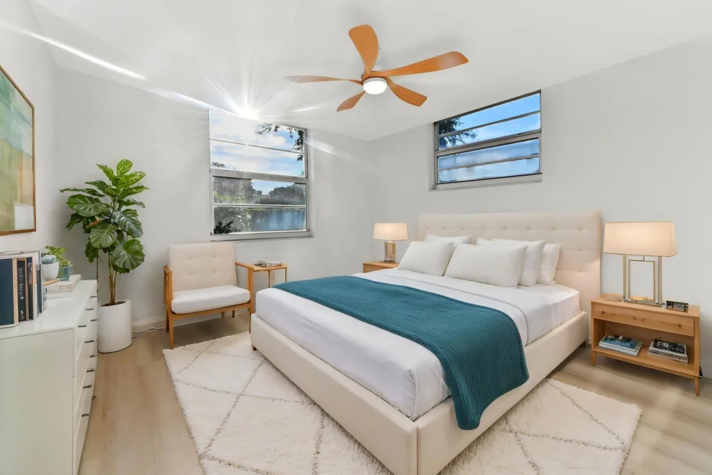 20261002165423459406000000-o - Lake Worth Homes - Lake Worth Homes Bright minimalist master bedroom with a white upholstered bed, teal throw, two wooden nightstands, and a potted plant by the dresser.
