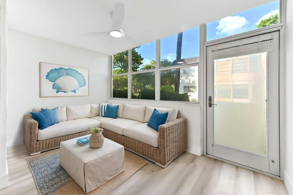 20261002165419383826000000-o - Lake Worth Homes - Lake Worth Homes Bright sunroom with a tan wicker L-shaped sofa, cream cushions, and blue accent pillows; coastal shell artwork on white wall and large window wall behind.