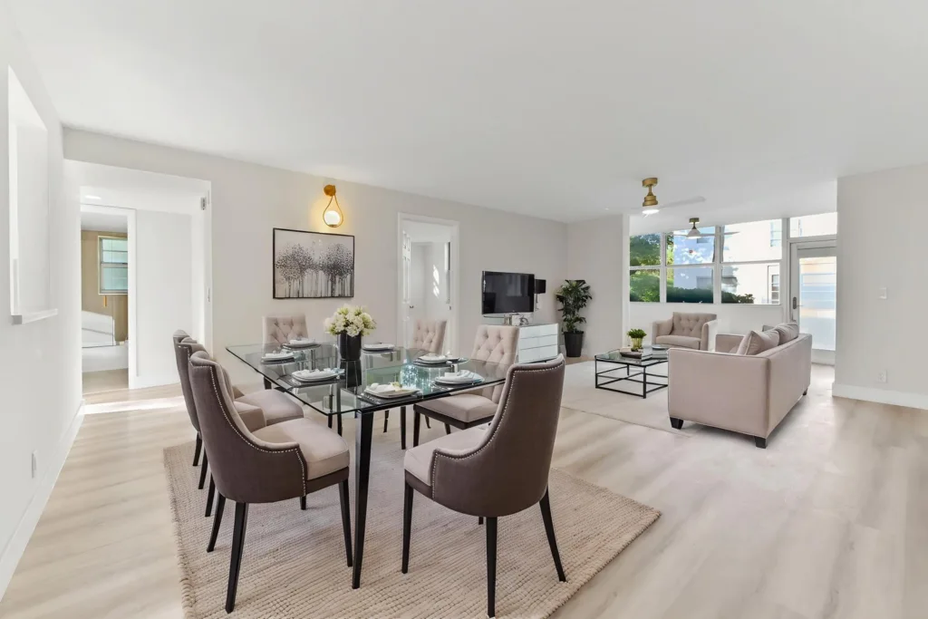 20261002165414314360000000-o - Lake Worth Homes - Lake Worth Homes Open-plan living and dining space with a glass dining table and six beige upholstered chairs, a beige sofa and armchairs, a wall-mounted TV, and large windows filling the room with light.