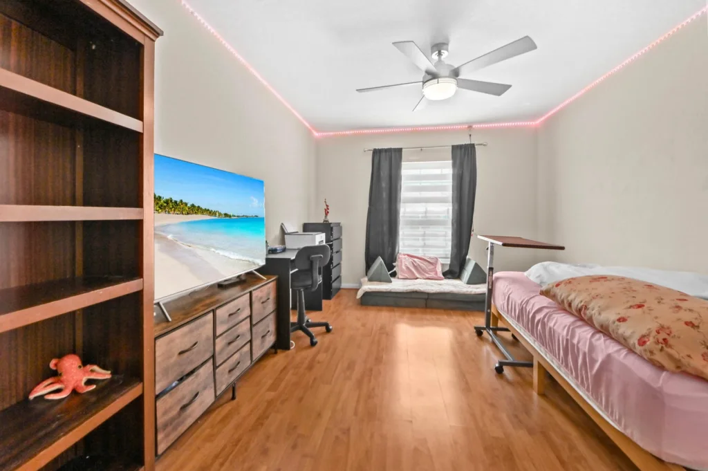 Bedroom with a workspace, TV on a dresser, and a bed; pink LED strips along the ceiling corner, wooden floor, and a window with dark curtains.