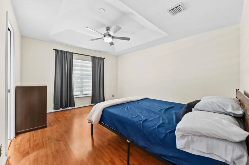 Bedroom with a low platform bed in navy sheets and gray pillows, hardwood floors, and dark curtains under a ceiling fan.