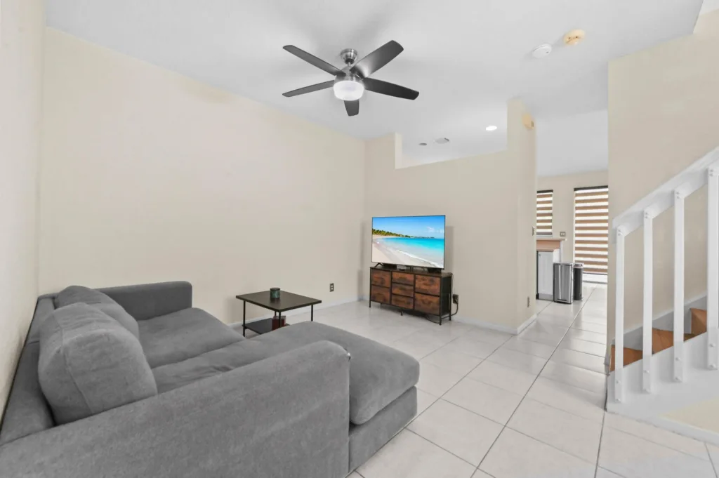 Living room with a gray sectional sofa facing a TV on a wooden stand, a small coffee table, and white tiled floor; stairs rise on the right side of the room.