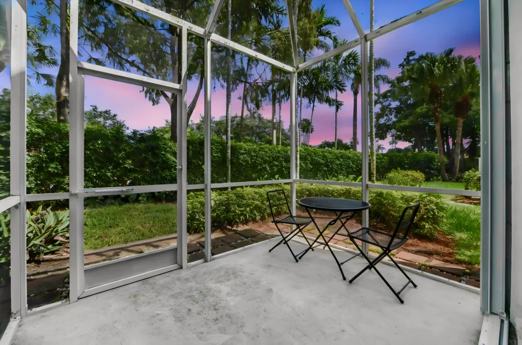Screened patio with a round black table and two folding chairs, overlooking a green garden with palm trees at sunset.
