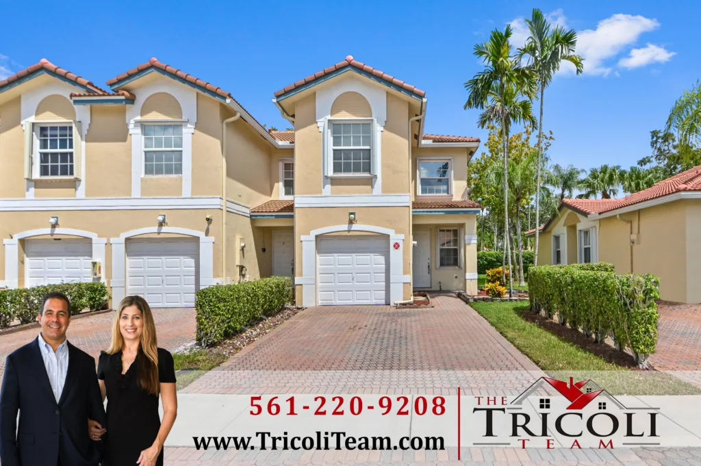 Smiling real estate agents stand in front of beige townhouses with a promotional banner for The Tricoli Team and a phone number.