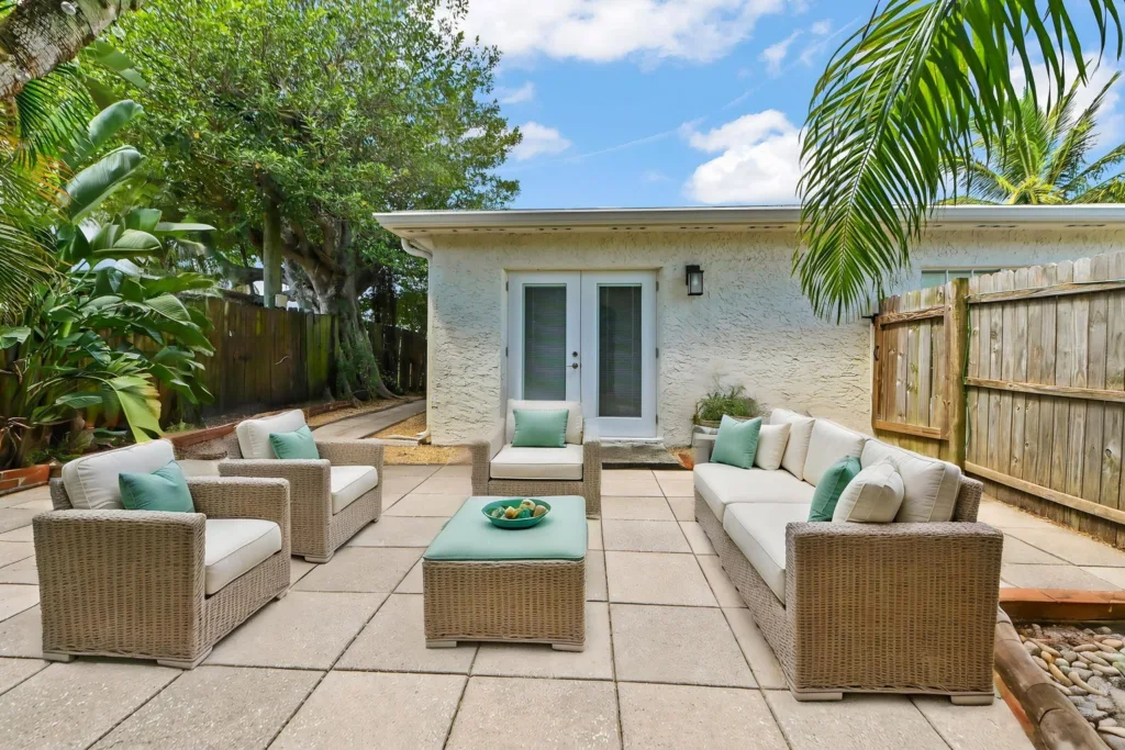 Patio with beige wicker seating and teal cushions arranged around a matching coffee table with a bowl of fruit, facing a white French door, surrounded by tropical plants.