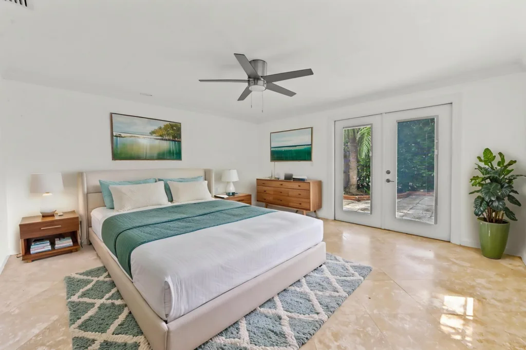 Bright modern bedroom with a beige upholstered bed, teal bedding, two wooden nightstands, and large glass French doors to a garden outside.